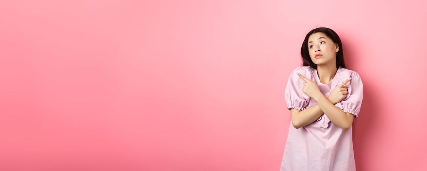 Indecisive asian woman cant decide, pointing fingers sideways and look pensive aside, making choice, standing confused on pink background.