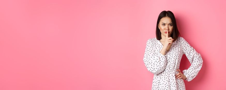 Angry asian female model shushing, frowning and hushing, showing taboo gesture, standing displeased against pink background, demand silence.