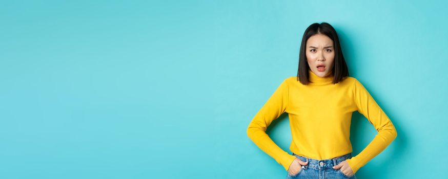 Portrait of disappointed korean woman in yellow sweater, frowning and looking upset, standing over blue background.