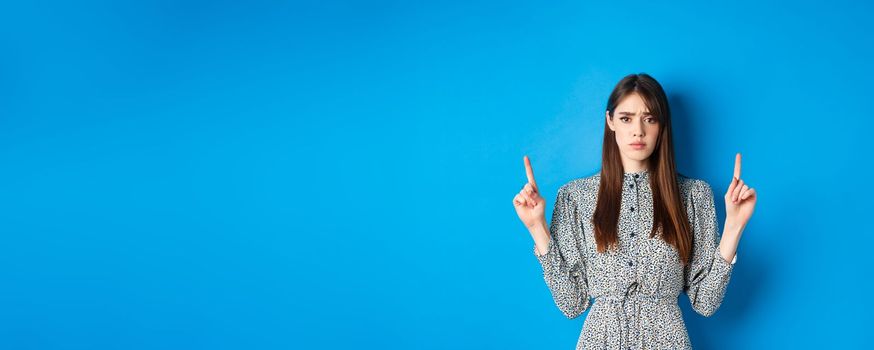 Hesitant and worried young woman frowning, pointing fingers up with sad face, standing on blue background.