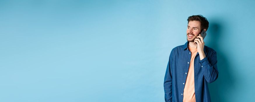 Handsome caucasian man talking on mobile phone, looking aside at empty space with carefree smile, standing on blue background.