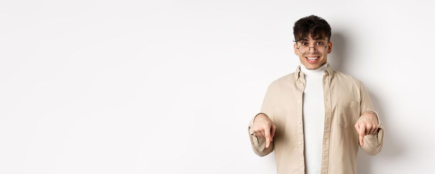 Portrait of happy young guy in glasses showing cool promo, pointing fingers down and smiling excited, standing on white background.