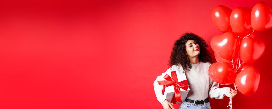 Romantic woman looking dreamy at heart balloons from lover, holding Valentines day gift in cute wrapped box, standing on red background.