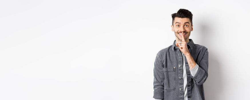 Happy guy shushing at camera, smiling with finger on lips, asking to keep quiet, telling big secret or making surprise, standing on white background.