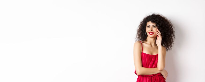 Elegant and romantic woman in red dress and makeup, smiling and touching her face, looking aside with happy expression, white background.