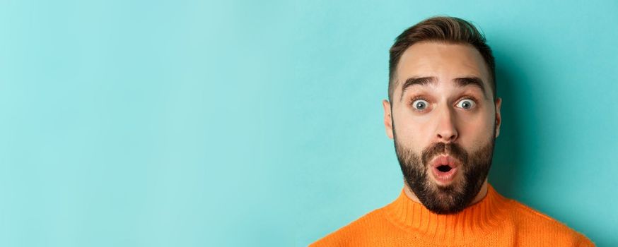 Headshot of handsome caucasian man with beard standing in orange sweater against turquoise background, saying wow and staring surprised at camera.