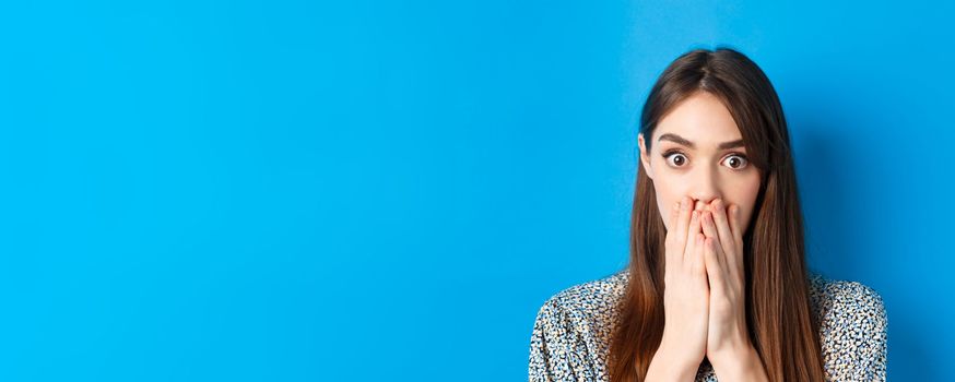 People emotions concept. Shocked young woman face, covering mouth with hands and gasping, look startled, standing on blue background.