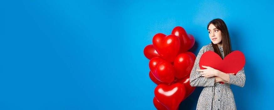 Valentines day. Beautiful and romantic woman looking pensive at balloons, hugging big red heart and smiling, waiting for love, blue background.