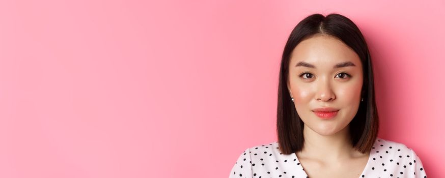 Beauty and skin care concept. Headshot of young feminine asian woman with short hairstyle, looking at camera, standing over pink background.