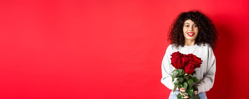 Romantic woman with curly hairstyle receive bouquet of red roses on Valentines day, looking tender at lover, standing on studio background.