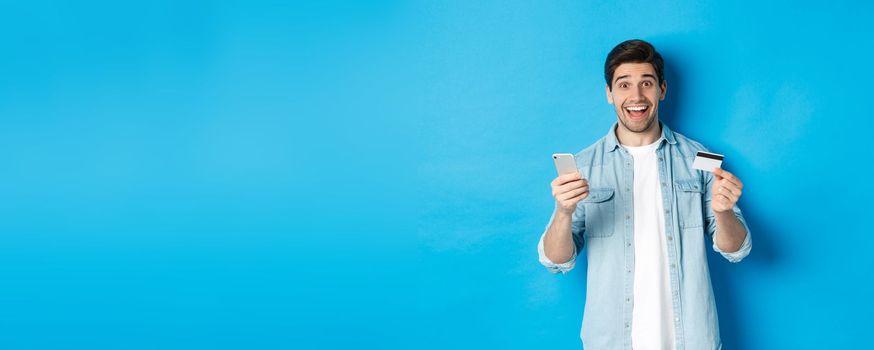 Amazed handsome man shopping online, holding mobile phone and credit card, smiling while paying for internet purchase, standing over blue background.