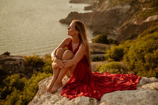 Sea woman sunset coast. Beautiful sensual woman in a long red dress and with long hair, sitting on a rock above the beautiful sea in a large bay