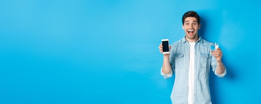 Happy man showing glass of water and mobile screen, recommending smartphone health app, standing over blue background.