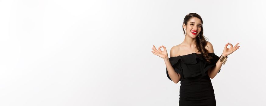 Fashion and beauty. Attractive brunette woman in black dress, showing okay signs and smiling satisfied, approve and recommend, standing over white background.