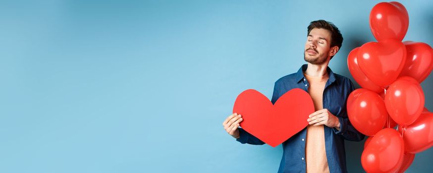 Valentines day and love concept. Dreamy man with closed eyes, holding romantic red heart cutout and standing near hearts balloons, blue background.