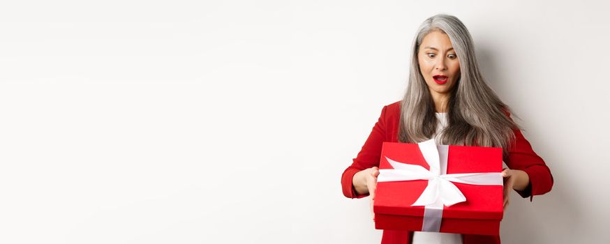 Surprised asian senior woman receiving present on mother day, holding red box with gift and looking amazed, white background.
