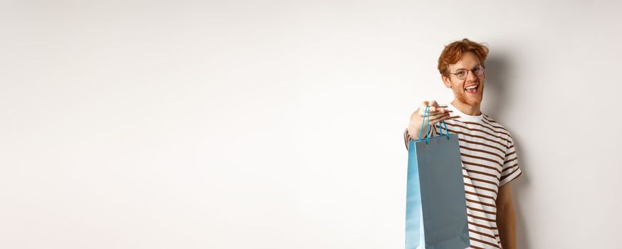 Funny handsome boyfriend giving shopping bag with gift, congratulate with valentines day and smiling, standing over white background.