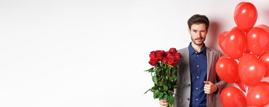 Confident boyfriend in suit going on romantic date, winking at camera, holding bouquet of red roses, standing near heart balloon and winking, standing over white background.