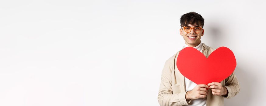 Handsome hipster guy with sunglasses and earring, waiting for true love on Valentines day, holding big red heart and smiling, standing over white background.