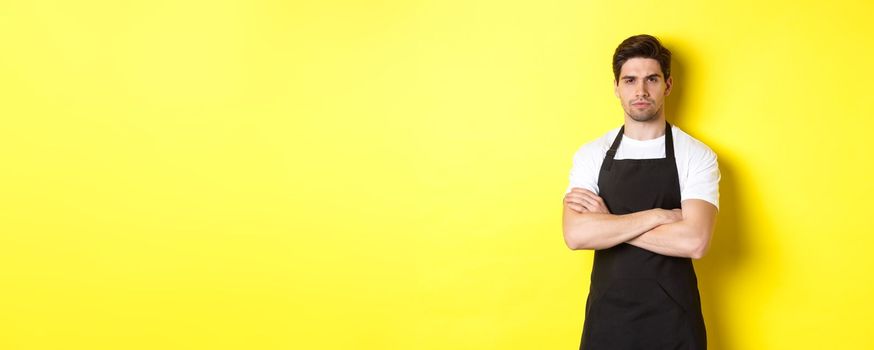 Suspicious barista squinting, cross arms on chest and looking at something with disbelief, standing over yellow background.