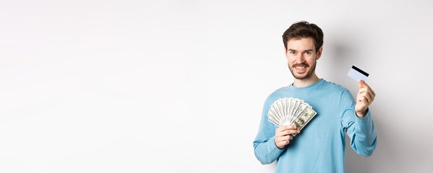 Handsome young man smiling and offering payment in cash and contactless, showing money with plastic credit card, standing on white background.