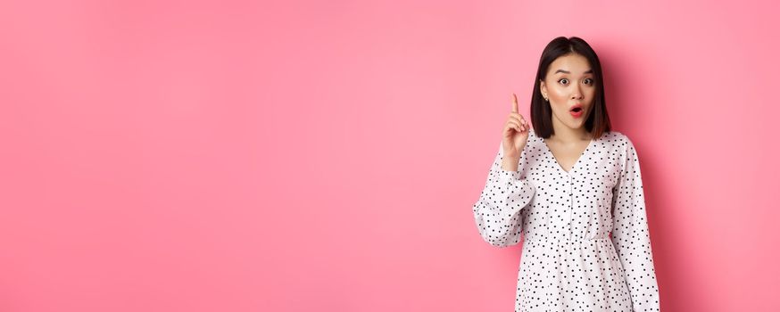 Excited asian girl having an idea, saying suggestion, raising finger and talking, standing over pink background.