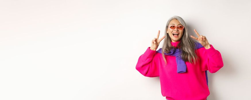 Fashion and beauty concept. Image of stylish asian senior woman in sunglasses smiling, showing peace signs and looking happy, standing over white background.