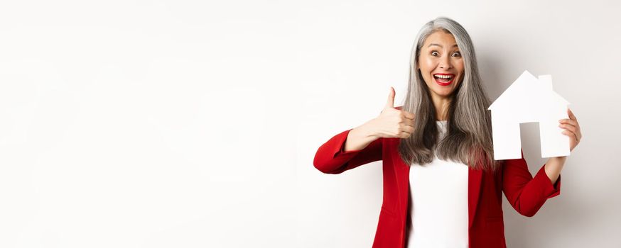 Real estate. Portrait of asian female broker showing thumb-up and paper house cutout, recommending agency to buy property, standing happy over white background.