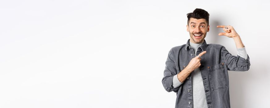 Excited man showing small size, pointing finger at little thing and smiling, standing against white background.
