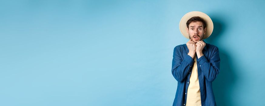 Scared young tourist trembling from fear, looking at something scary on summer vacation, wearing straw hat, standing on blue background.