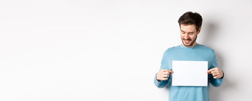 Young caucasian man looking at empty paper, reading logo, standing on white background.