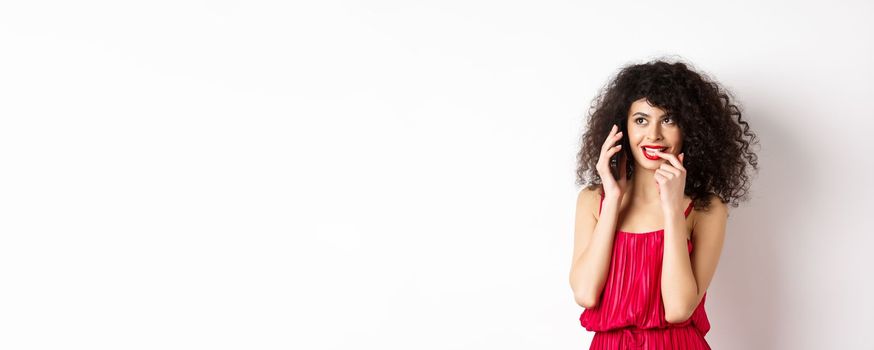 Silly fashionable woman in red dress and lips, biting fingernail duing phone call, thinking on mobile conversation, standing on white background.