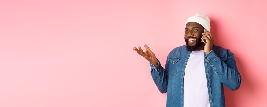 Handsome modern african-american man talking on mobile phone, smiling and discussing something, standing over pink background.