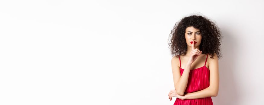 Angry elegant woman in red dress hushing and frowning, tell to be quiet, asking for silence, standing over white background.