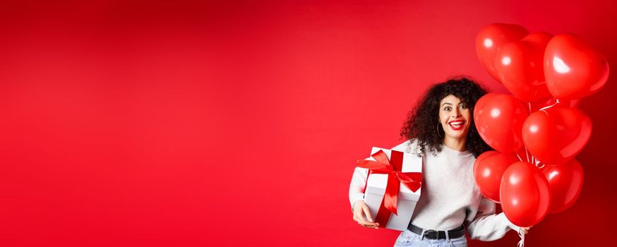 Holidays and celebration. Happy birthday girl holding gift and posing near party helium balloons, smiling excited at camera, red background.