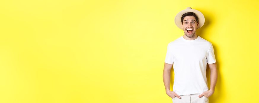 Surprised man tourist in straw hat looking happy, react amazed to travel agency advertisement, standing over yellow background.