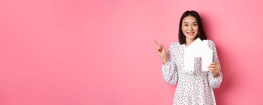 Real estate. Excited asian woman showing paper house model, pointing left at copy space, standing over pink background.