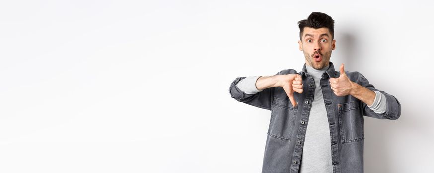 Excited guy show thumbs up and down judging and making decision, rate something interesting, standing on white background.