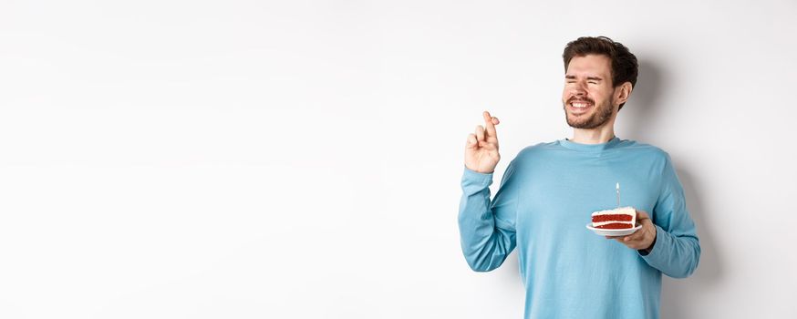 Celebration and holidays concept. Young man celebrating birthday, cross fingers for good luck, making wish on bday cake with lit candle, standing over white background.