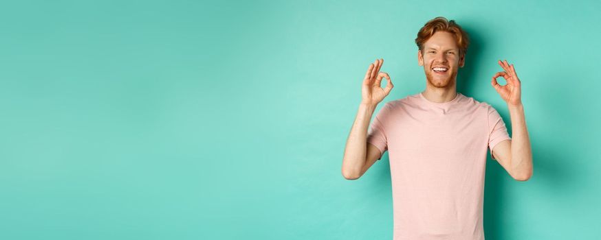 Attractive young man in t-shirt smiling satisfied, nod in approval and showing OK sign, approve and agree with something cool, standing over turquoise background.