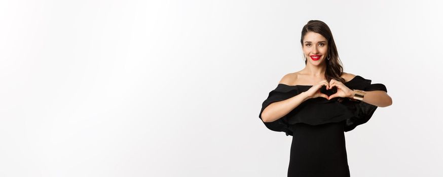 Fashion and beauty. Beautiful young bruentte female showing heart sign and smiling, express love and sympathy, wearing black dress, white background.