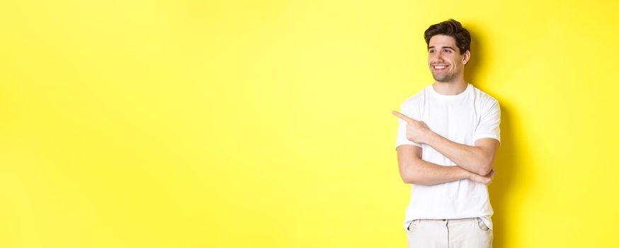 Handsome smiling man in white clothes, looking and pointing finger left at banner, standing over yellow background.