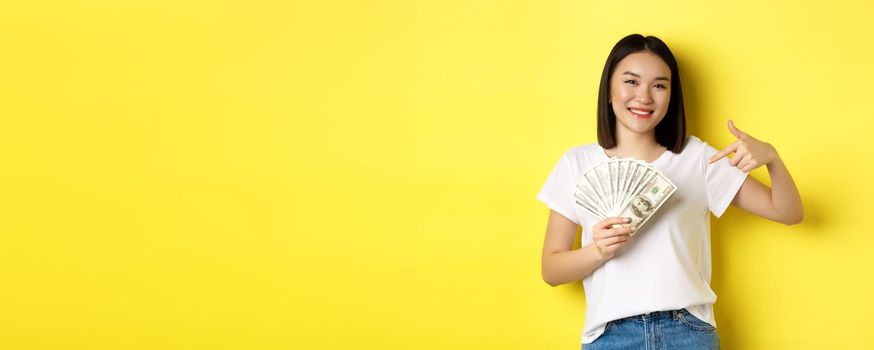 Young asian woman smiling, showing prize money, pointing finger at dollars, standing over yellow background.