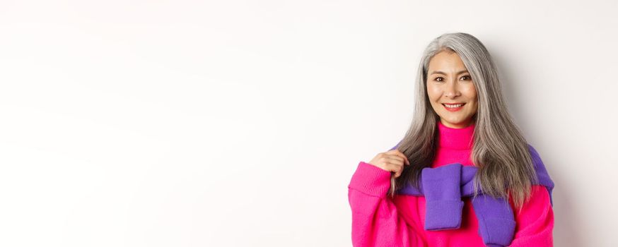 Close-up of beautiful asian senior woman in trendy pink sweater, smiling cheerful at camera, standing over white background.