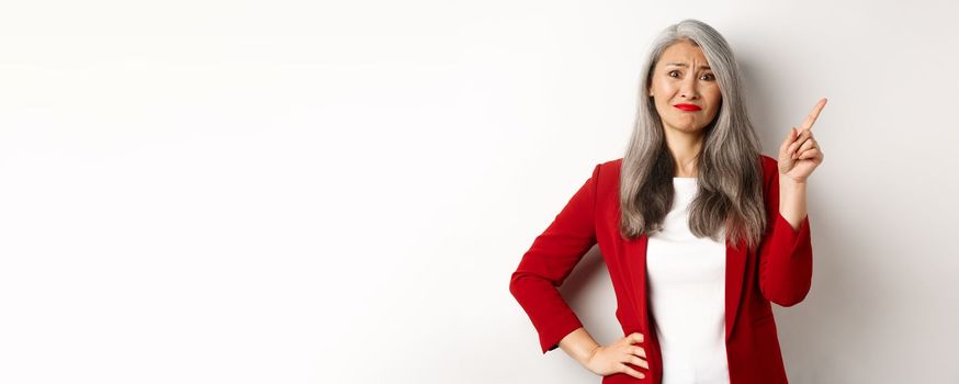 Disappointed and skeptical asian female entrepreneur pointing finger upper right corner, grimacing and frowning upset, showing bad promo, white background.