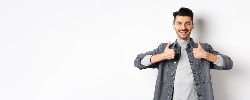 Handsome smiling man showing thumbs-up in approval, praise good work, satisfied with nice choice, recommending something cool, standing on white background.