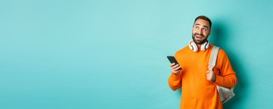 Image of handsome young man with backpack and headphones, imaging or thinking while using smartphone, standing over light blue background.