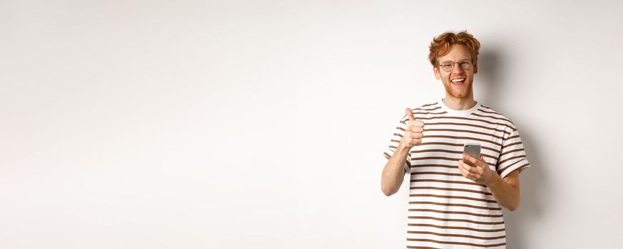 Technology and e-commerce concept. Satisfied male model with red hair, showing thumbs-up and holding smartphone, white background.