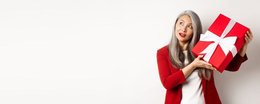 Elegant asian senior woman shaking gift box, trying to guess what inside present, standing over white background. Holidays and celebration concept.