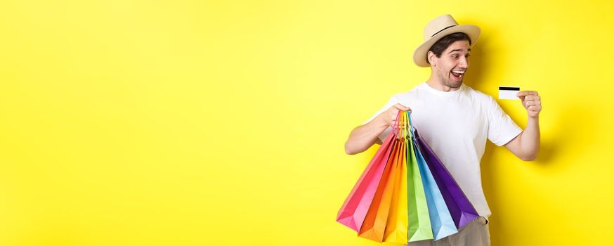 Excited man going shopping on vacation, looking satisfied at credit card, holding paper bags, standing over yellow background.